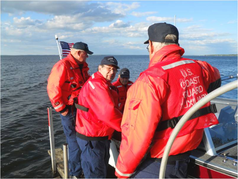 Three men in a boat and two on the dock, they are all wearing orange life jackets, at the dock preparing to go out on a boat