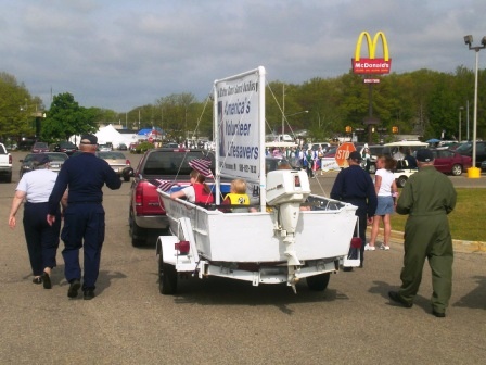 Several people in Coast Guard Uniforms walking along a parade route with a Auxiliary Decorated boat parade